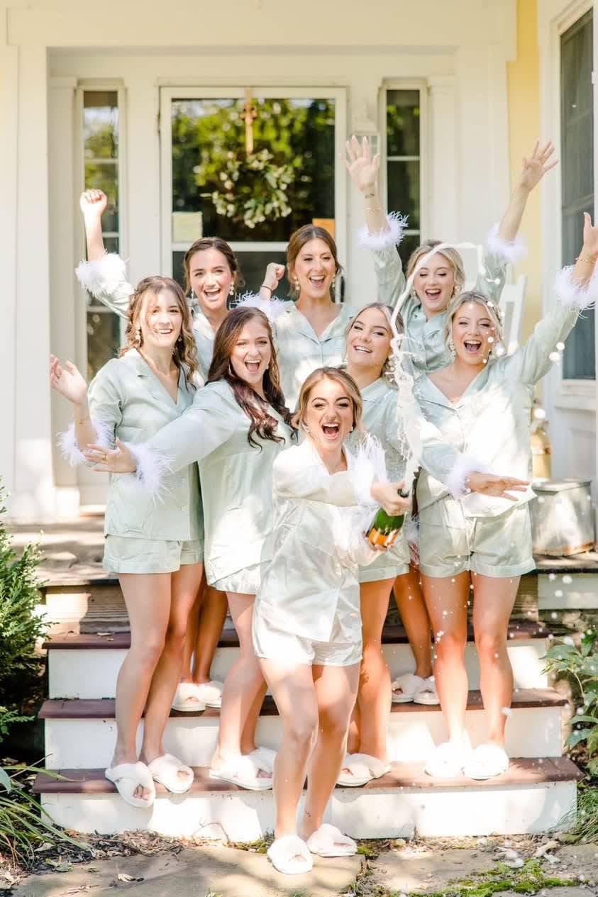 A group of women in matching sage green pyjama sets on a white porch, arms up, one of them spraying a bottle of champagne