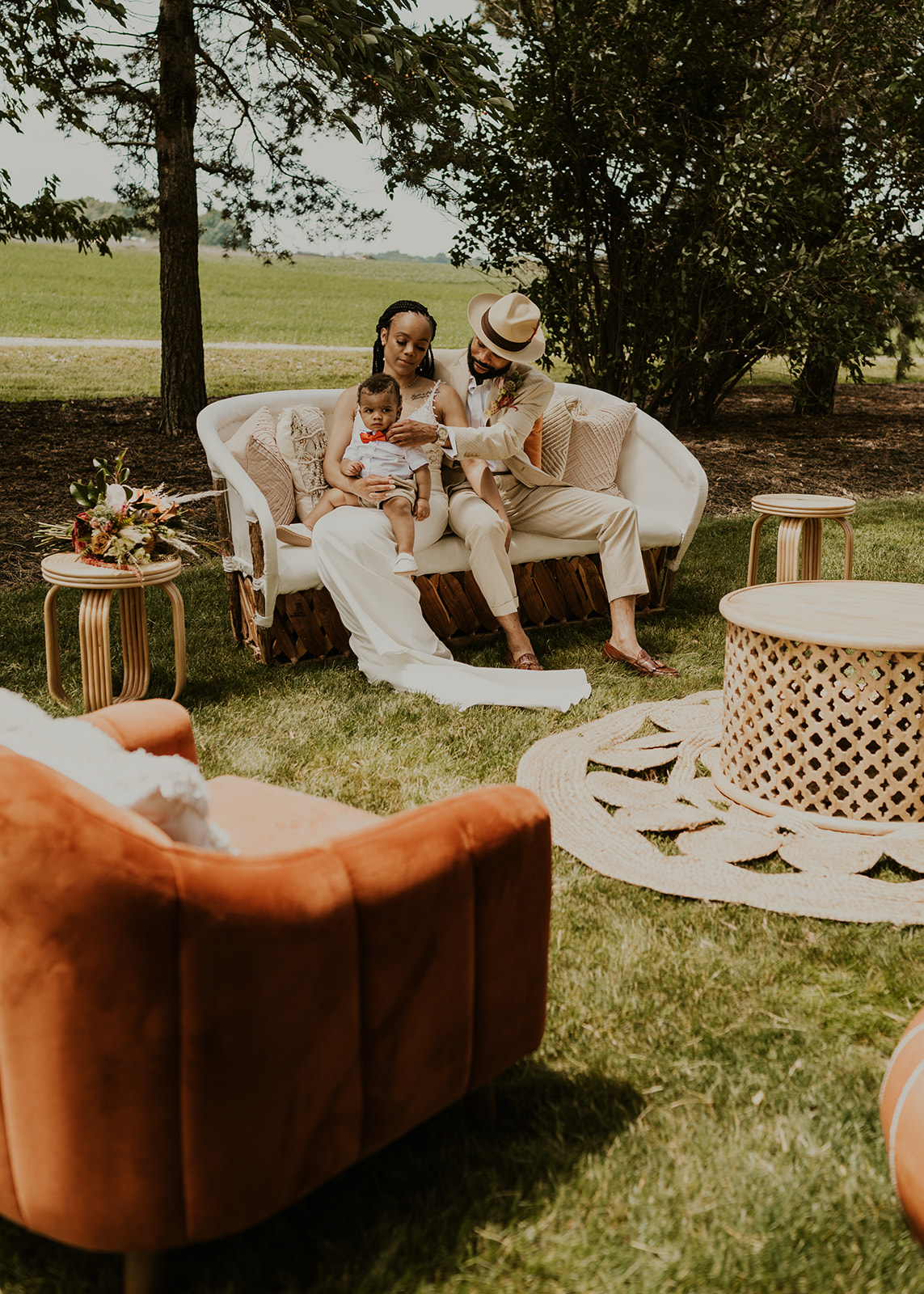 An outdoor lounge set up on the lawn at RW How: a cream rattan sofa and a rust velvet armchair on a woven rug under the trees, with the couple and their baby sitting together