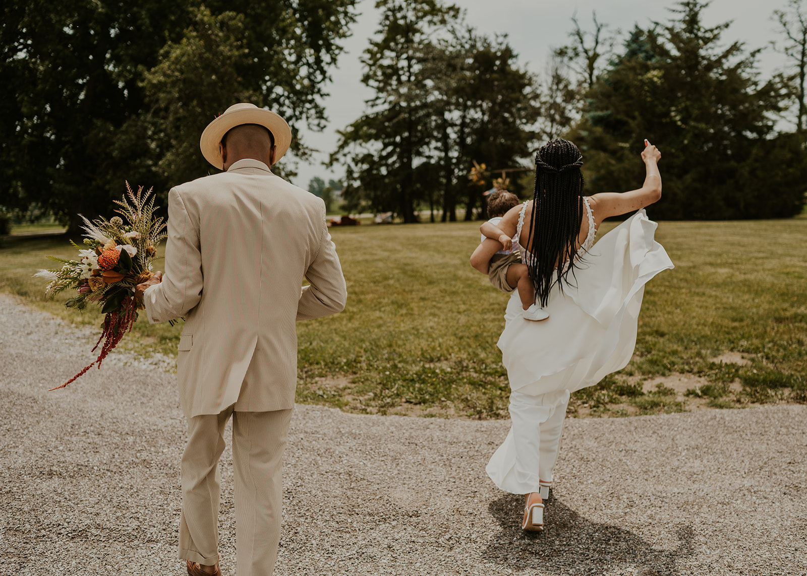 A couple walking away down the gravel drive at RW How after their ceremony, him in a tan suit and straw hat carrying the bouquet, her carrying their toddler with her dress flying out behind her