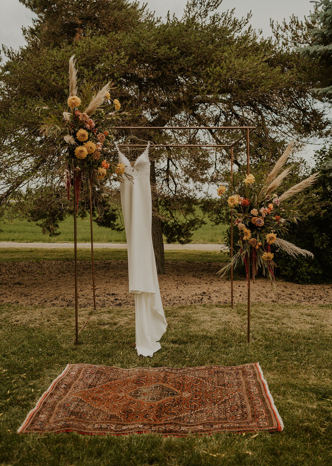 A wedding dress hanging from a copper arbour dressed with pampas grass and rust dahlias, a vintage Persian rug on the grass beneath