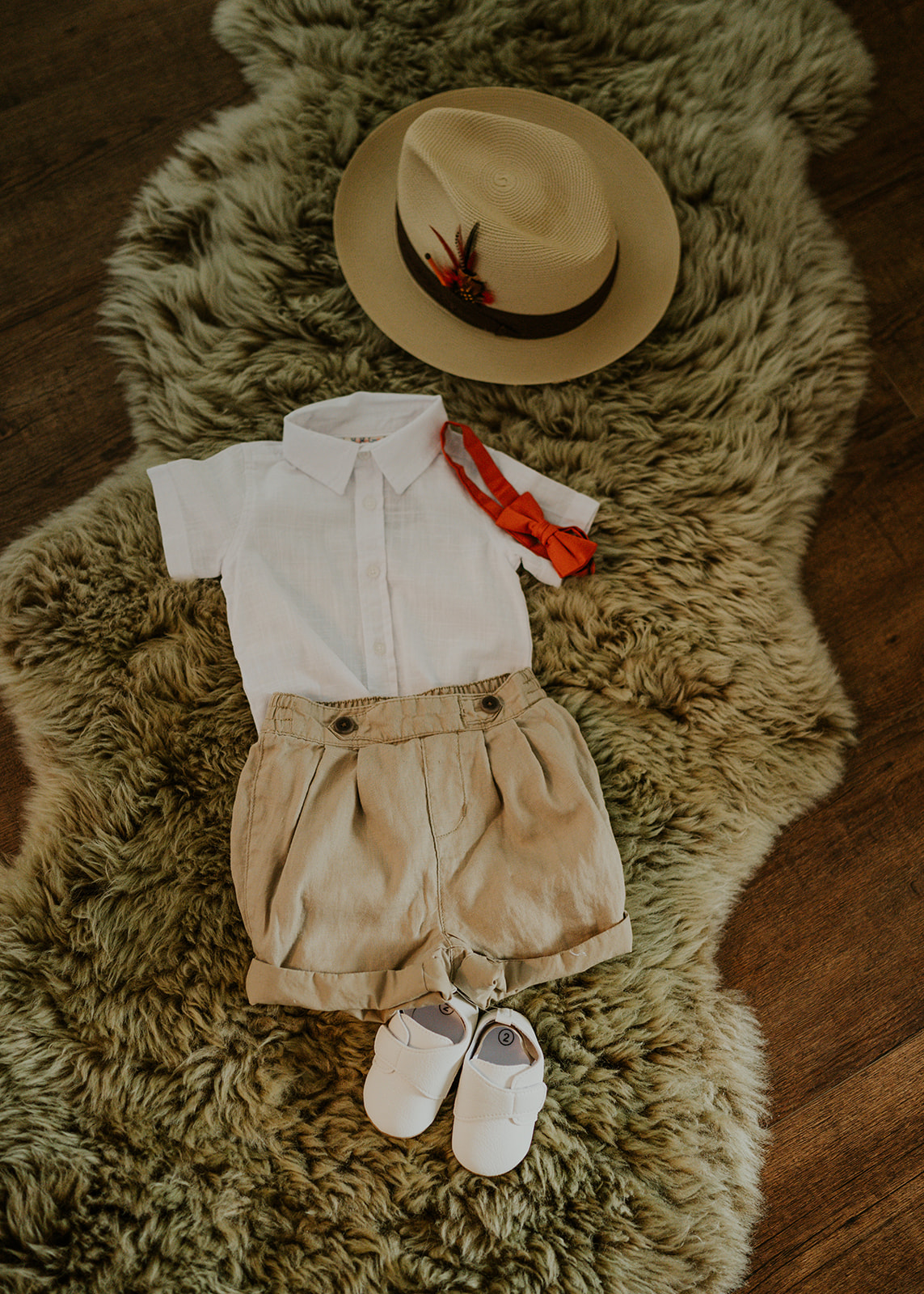 A straw hat, a small linen shirt with a rust bow tie, tan shorts and white shoes laid out on a green sheepskin
