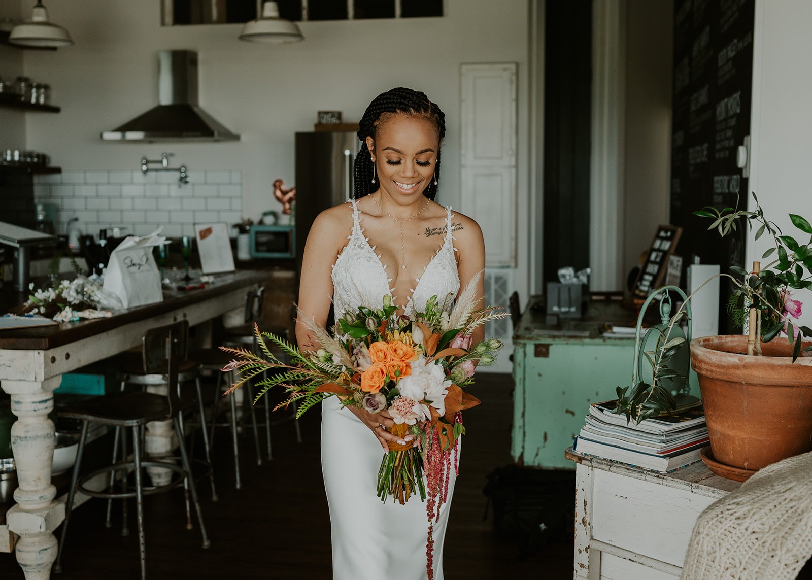 A bride holding a bouquet of orange roses, pampas grass and trailing amaranthus in a farmhouse kitchen with white subway tile and a chalkboard menu behind her