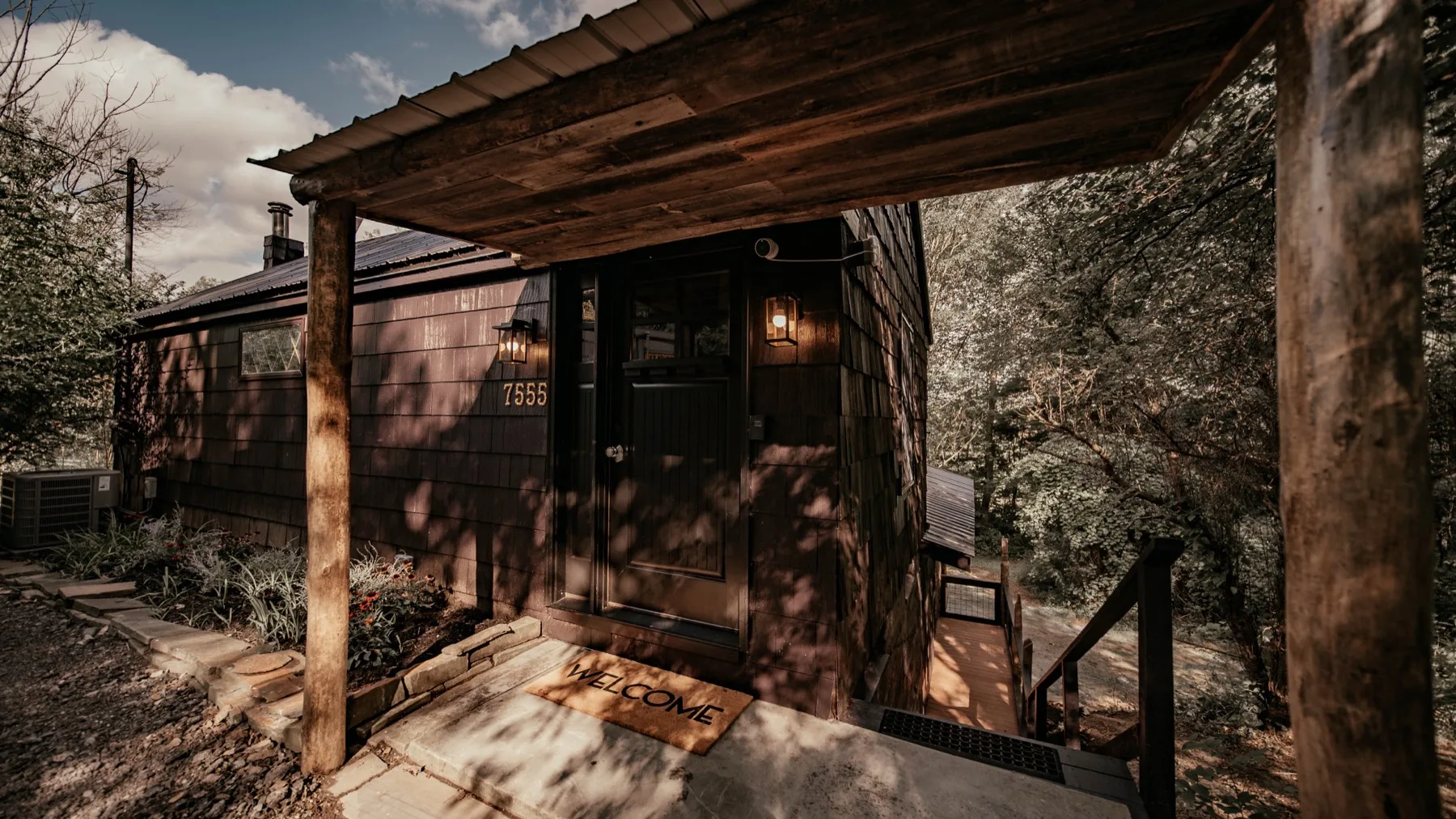 The cabin front door at dusk, house number 7555 above a welcome mat with the sconces lit on cedar shingle siding