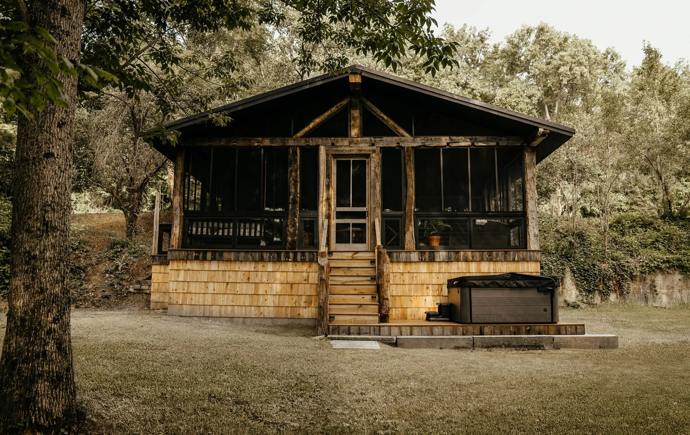 The 19A Cabin seen from the lawn, a cedar-shingle cabin with a black-roofed screened porch under tree canopy