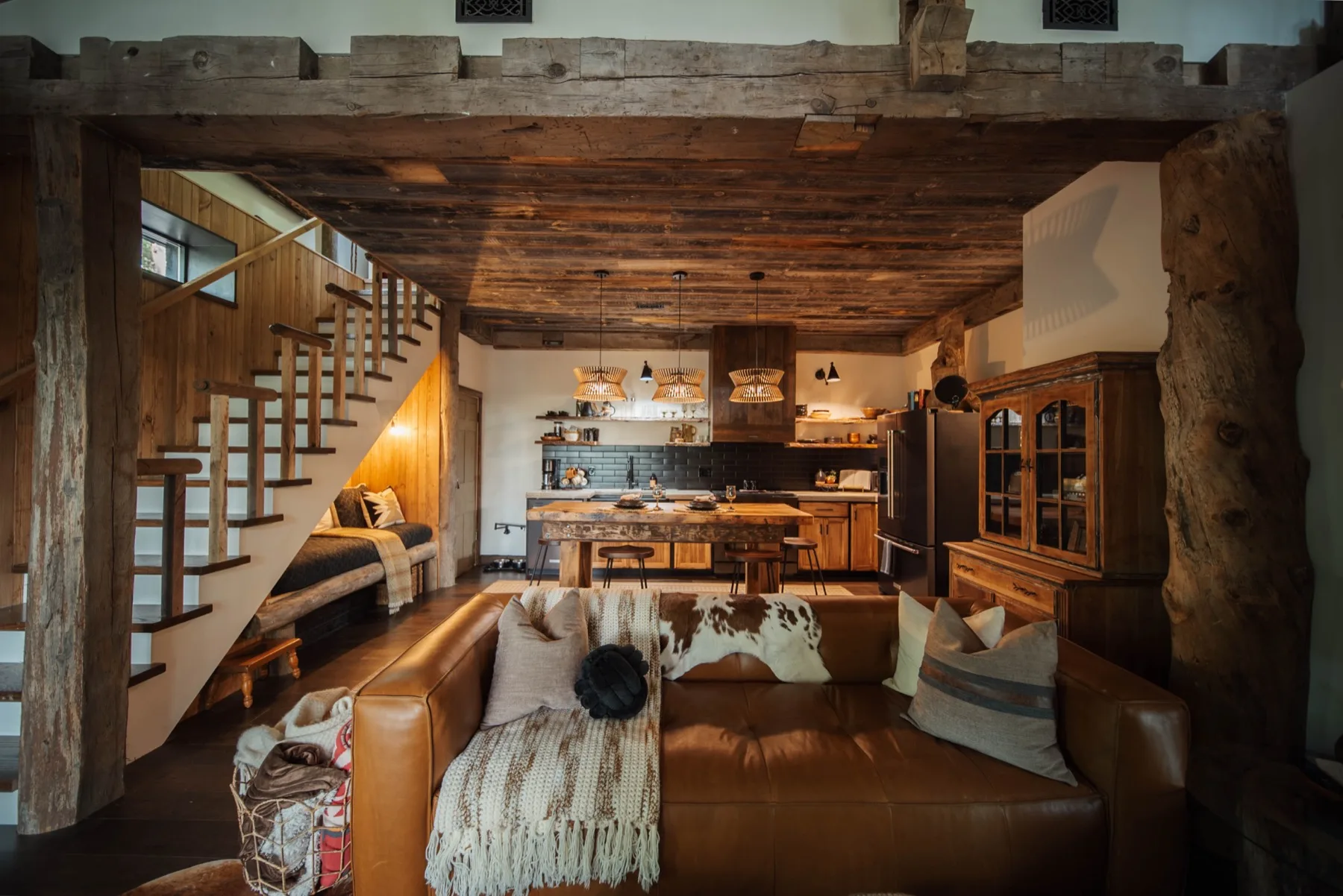 Open living area under hand-hewn reclaimed beams, a tan leather sectional and an open stair up to the loft