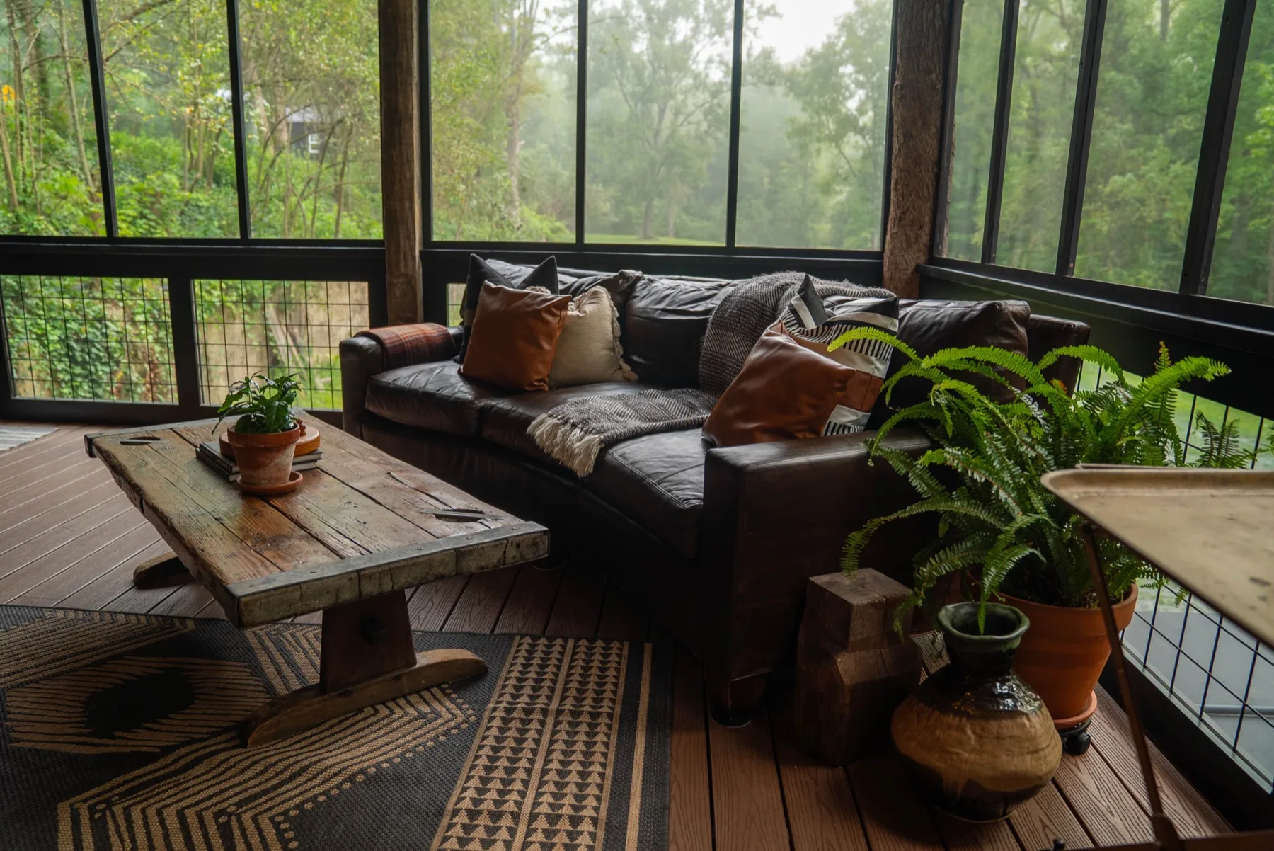 A curved dark leather sofa in the corner of the screened porch, facing a misty view of the woods