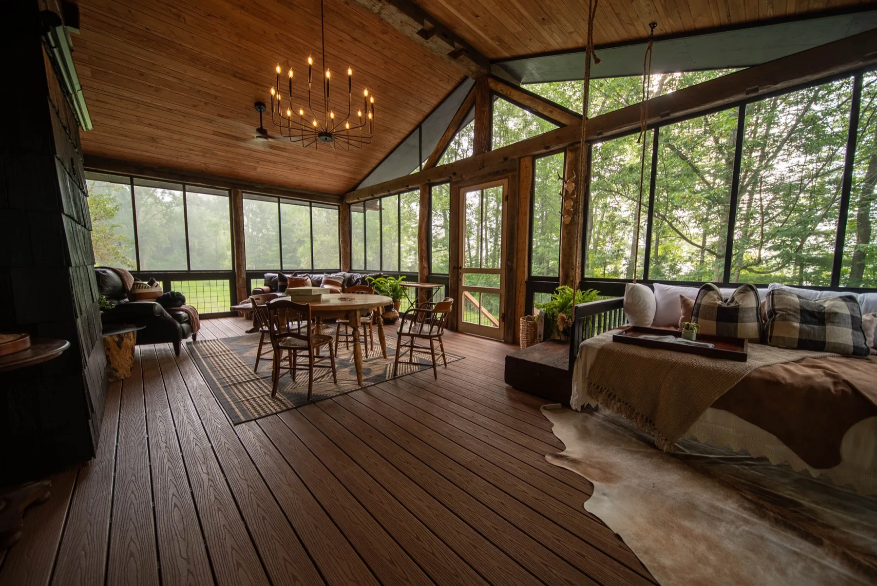 The full-width screened porch at the 19A Cabin, vaulted pine ceiling and a wagon-wheel chandelier over a farm table, with green woods through the screens