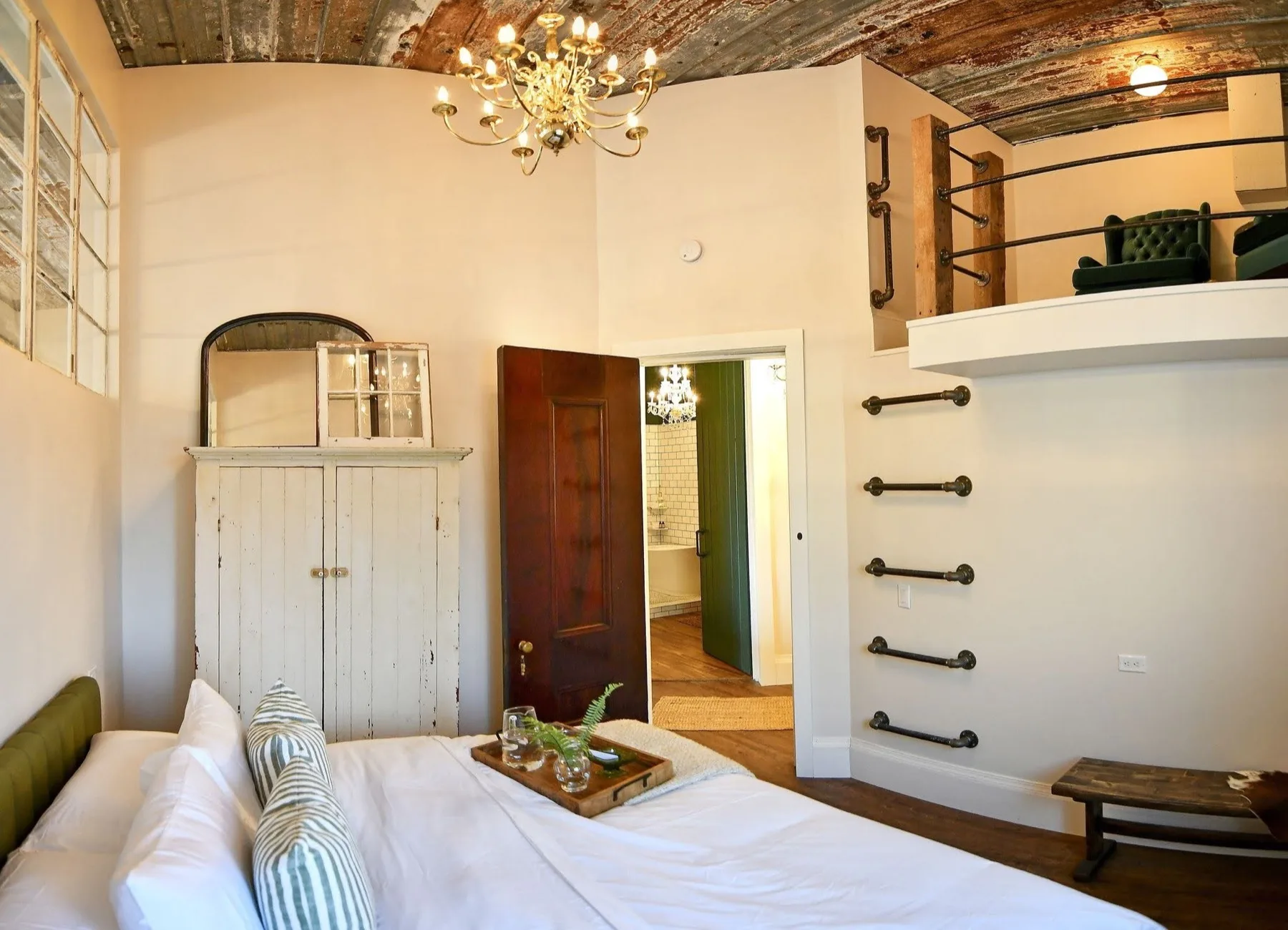 King bedroom under a barrel-vaulted tin ceiling with a brass chandelier, the ensuite door open to a green tiled bath