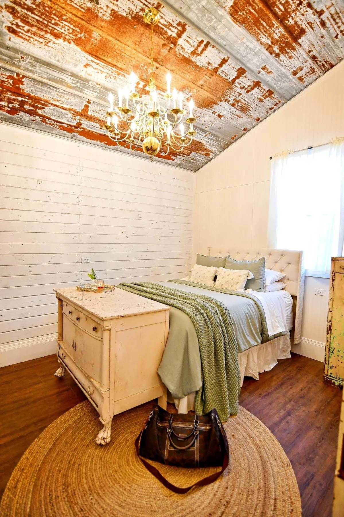 Second bedroom under weathered rust-coloured tin, with a brass chandelier and a distressed cream dresser