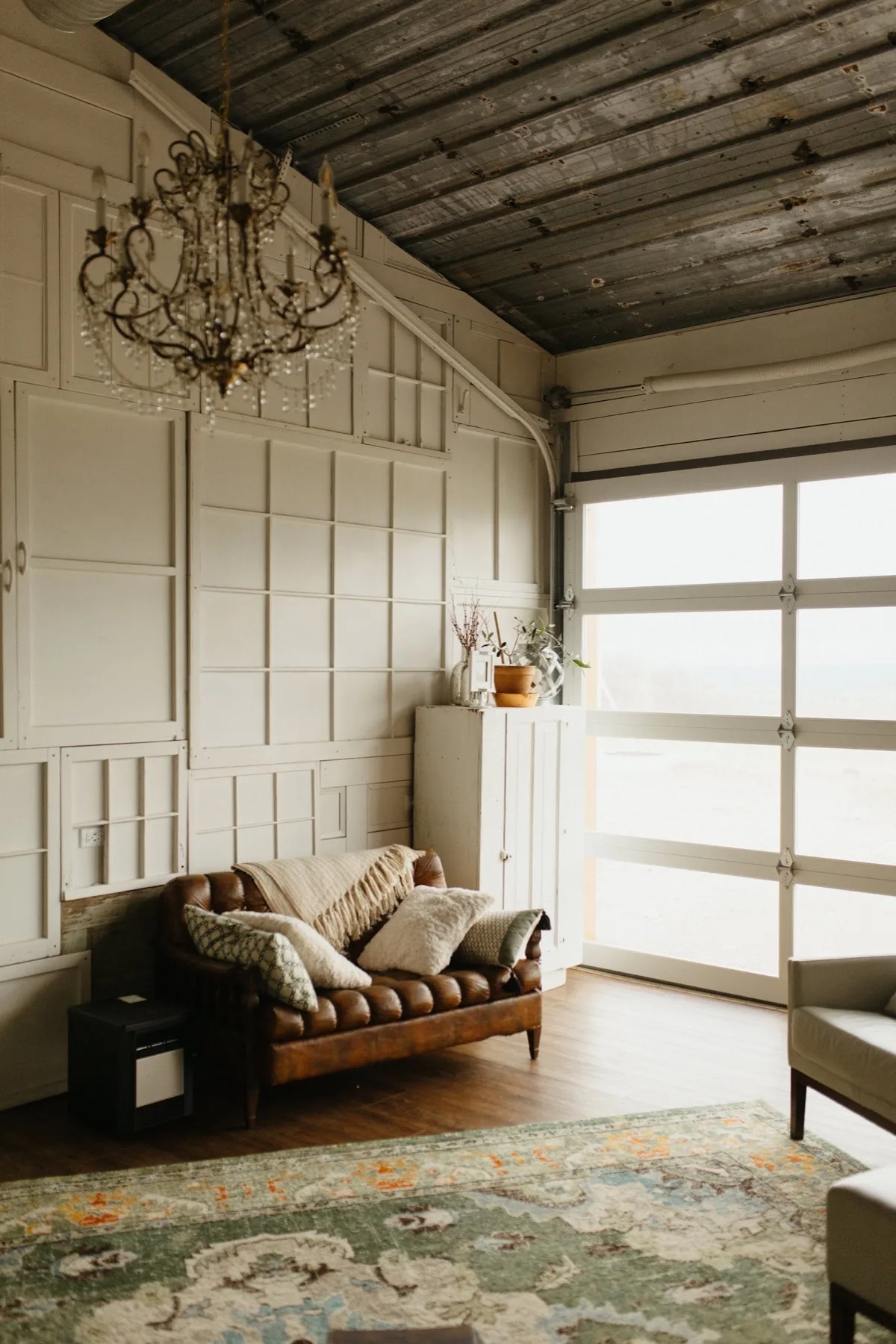 A brown leather loveseat with green pillows in the corner of the Highbanks living room, under a full-height glass garage door