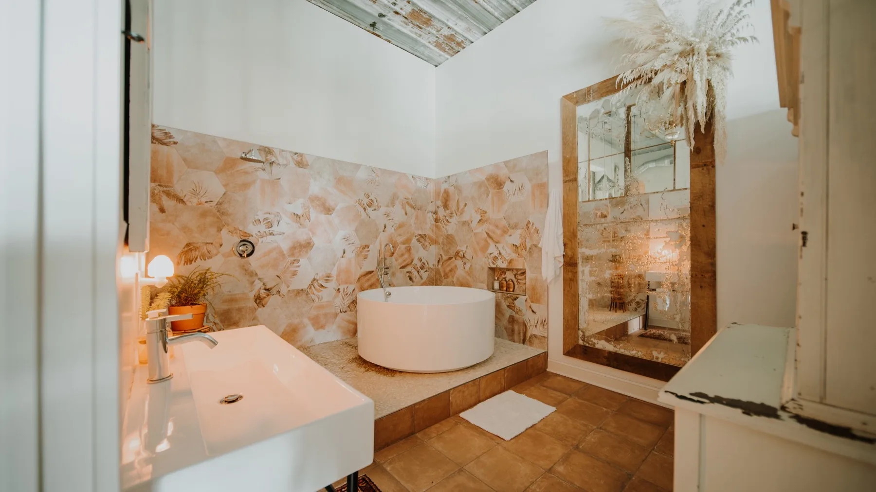 A round soaking tub on terracotta tile against patterned stone-look walls, beside an antique mirror dressed with dried pampas