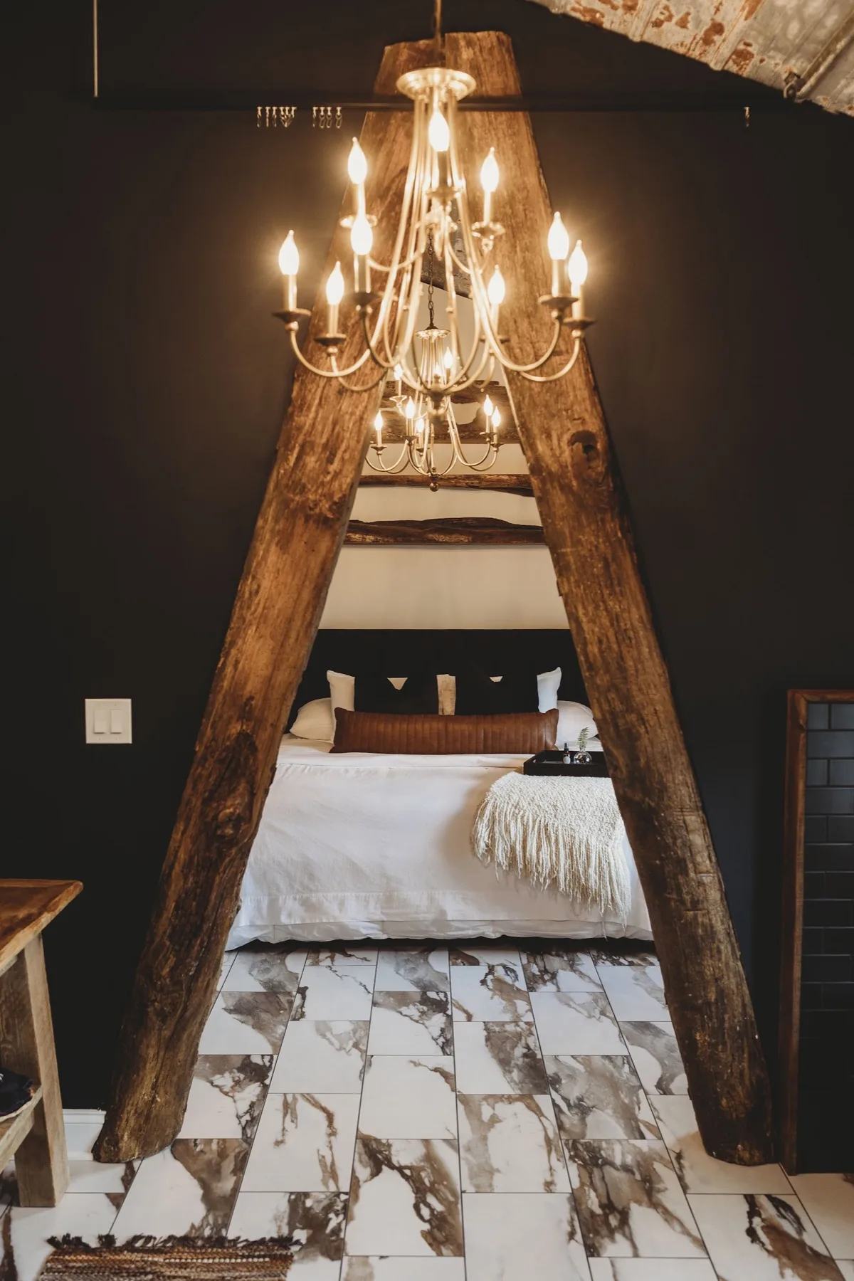 A reclaimed hand-hewn timber archway hung with a chandelier, framing the bed beyond it against black walls