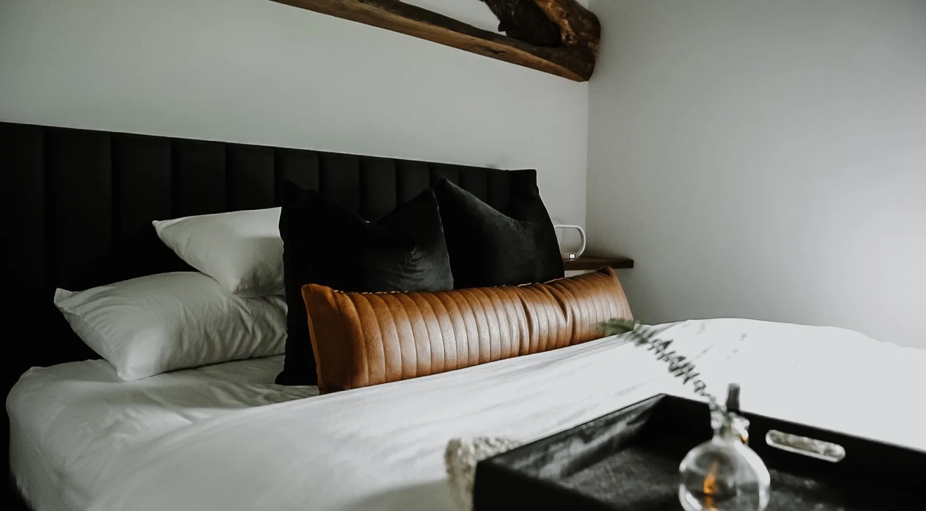 Bedroom with a black channel tufted headboard, a tan leather bolster and a reclaimed timber mantle shelf