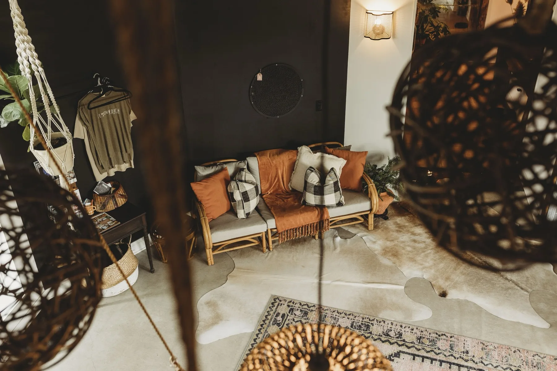 A dark boho nook with a rust and rattan loveseat, buffalo check pillows, a cowhide rug and macrame hangers