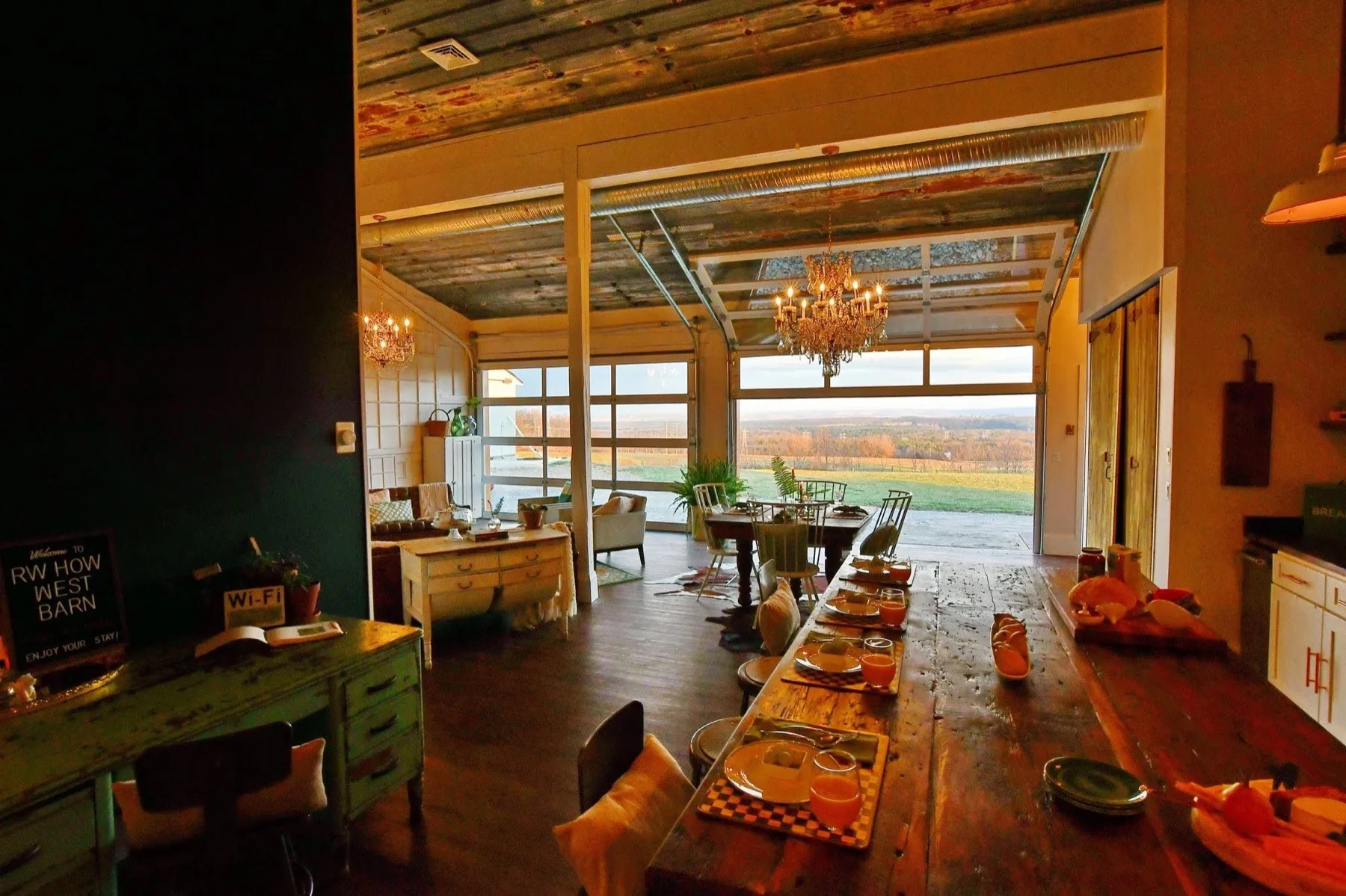 The West Barn great room at golden hour, a long reclaimed-wood table set for breakfast under crystal chandeliers with the glass garage doors rolled open onto the valley